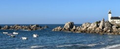 A white lighthouse stands on a granite rock outcrop at the edge of a calm bay in Kerlouan, Brittany. Small boats are anchored in the water, with a sandy beach in the foreground and clear blue sky above.