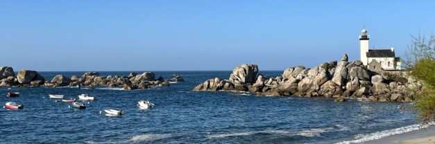 A white lighthouse stands on a granite rock outcrop at the edge of a calm bay in Kerlouan, Brittany. Small boats are anchored in the water, with a sandy beach in the foreground and clear blue sky above.