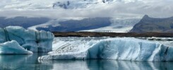 Chunks of blue-white glacier ice float in a lagoon in Iceland. Behind them, a much larger glacier and snow-capped mountains are mostly hidden by low cloud.
