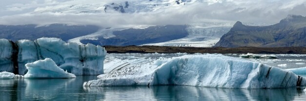 Chunks of blue-white glacier ice float in a lagoon in Iceland. Behind them, a much larger glacier and snow-capped mountains are mostly hidden by low cloud.