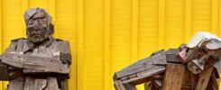 Two weathered wooden sculptures sitting on an outdoor bench against a bright yellow wall in Siglufjörður, Iceland. The left figure sits upright with arms crossed. The right figure is slumped forward, head resting near its knees."