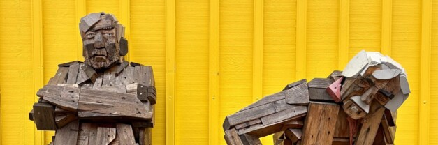 Two weathered wooden sculptures sitting on an outdoor bench against a bright yellow wall in Siglufjörður, Iceland. The left figure sits upright with arms crossed. The right figure is slumped forward, head resting near its knees."